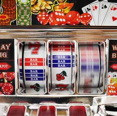 A close-up view of a slot machine with vibrant colors and various gambling symbols such as dice, coins, and playing cards. The reels display a combination of symbols, including bars, cherries, and the number seven. The chrome exterior and colorful details convey the theme of casino and gambling.