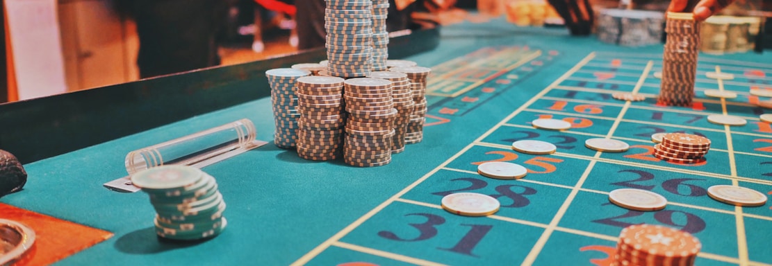 A casino scene with a roulette table features prominently, covered in green felt with colorful numbers and betting squares. Several stacks of poker chips are placed on different numbers, and the hands of several people are visible as they interact with the table. In the background, there are slot machines and more people, creating a vibrant and busy atmosphere.
