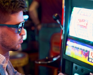 A man wearing sunglasses is intently focused on a slot machine, with colorful lights and graphics illuminating the screen. The scene is set indoors, likely in a casino or gaming area. His attire includes a patterned shirt and a blazer.
