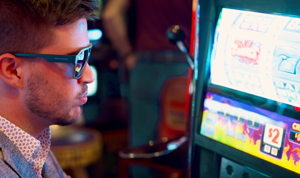 A man wearing sunglasses is intently focused on a slot machine, with colorful lights and graphics illuminating the screen. The scene is set indoors, likely in a casino or gaming area. His attire includes a patterned shirt and a blazer.
