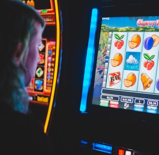 A person is closely observing a digital slot machine with colorful fruit symbols, bars, and a bell. The slot machine screen displays symbols like oranges, plums, cherries, and clouds. The background is dimly lit, creating a focused and somewhat intense atmosphere.