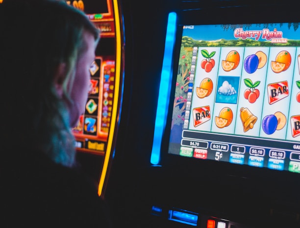 A person is closely observing a digital slot machine with colorful fruit symbols, bars, and a bell. The slot machine screen displays symbols like oranges, plums, cherries, and clouds. The background is dimly lit, creating a focused and somewhat intense atmosphere.