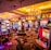 A vibrant casino interior filled with rows of colorful slot machines, each illuminated with bright lights and displays. The ceiling is adorned with elegant chandeliers and golden drapes, creating a luxurious atmosphere. The patterned carpet adds to the elaborate design of the space.