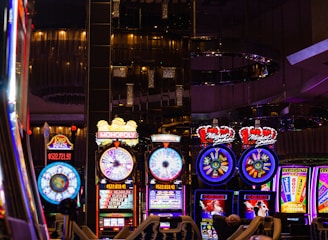 A lively casino scene featuring a row of bright, colorful slot machines with themes like Monopoly, Michael Jackson, and Lord of the Rings. The gaming machines display a variety of flashing lights and digital screens, creating an energetic and exciting atmosphere. Reflections of the lights are visible in glossy surfaces above the machines.