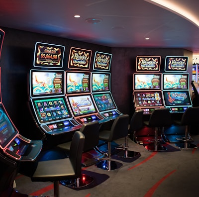 A row of brightly lit slot machines in a casino setting, each with colorful displays and various game themes. The machines are lined up against a dark wall and are unoccupied, with cushioned chairs placed in front of them. The area is dimly lit except for the glow from the screens, creating a vibrant and engaging atmosphere.