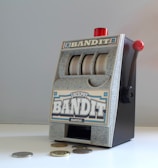 A small, vintage-style slot machine with the word 'Bandit' prominently displayed. It features a lever on the side with a red knob and three reels with a generic design. Several coins are scattered on the white surface in front of it.